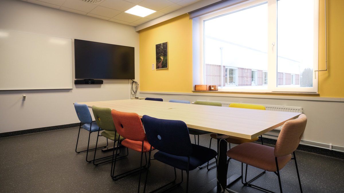 Ett mindre mötesrum med ett rektangulärt bord, färgglada stolar, en stor skärm och ett stort fönster som släpper in dagsljus.