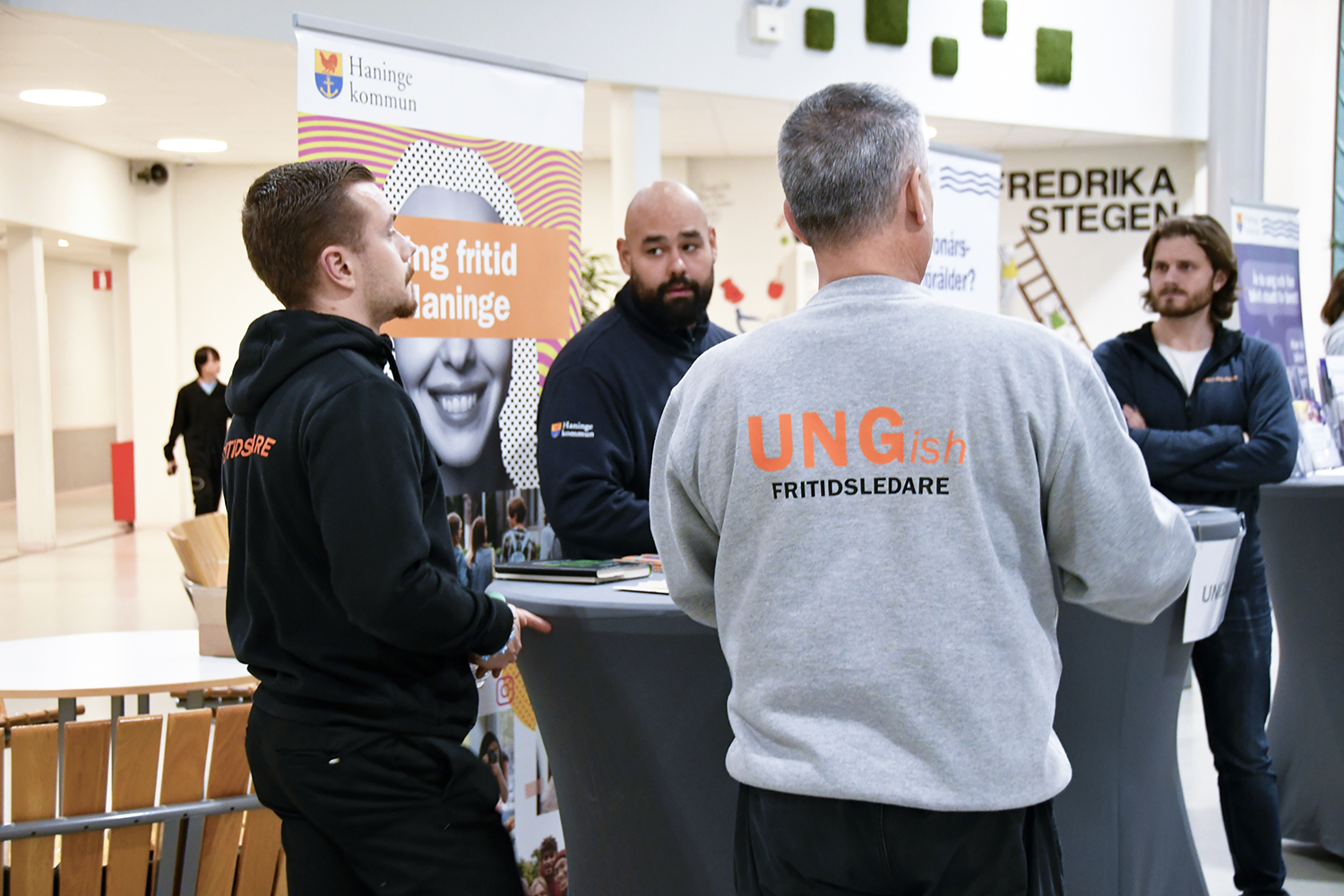 Grupp med ungdomsledare i tröjor märkta 'UNGish FRITIDSLEDARE' står vid informationsbord.