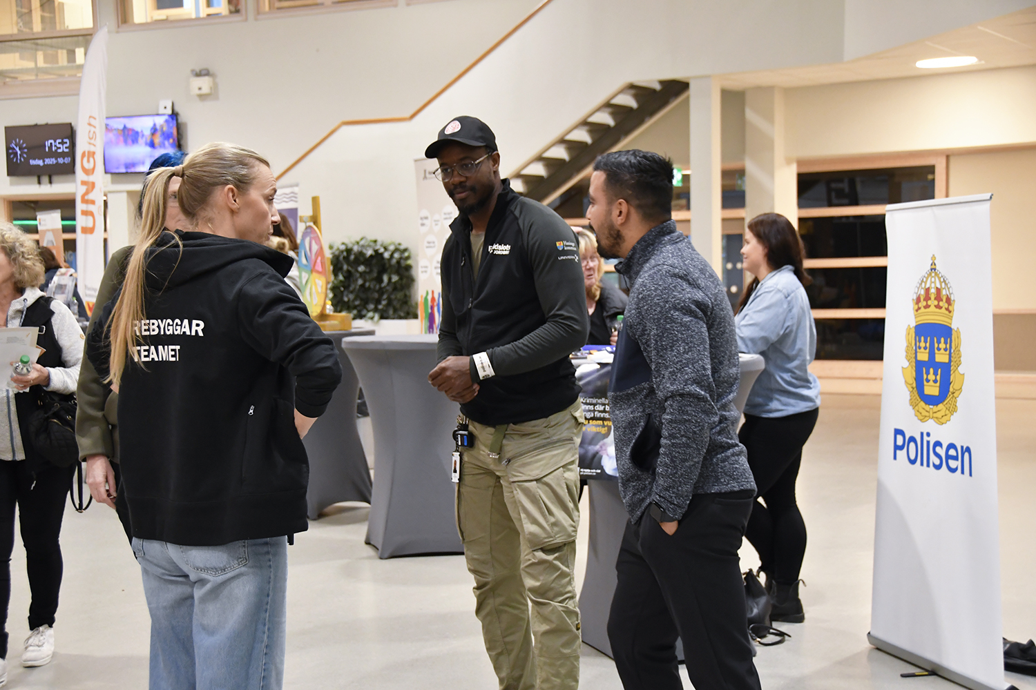 Tre personer samtalar vid ett evenemang, varav en bär jacka med texten "Förebyggarteamet" och en banner från Polisen syns i bakgrunden.