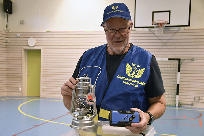 En representant från Civilförsvarsförbundet håller upp en oljelampa och en annan krispryl, iförd blå väst med texten 'www.civil.se'.