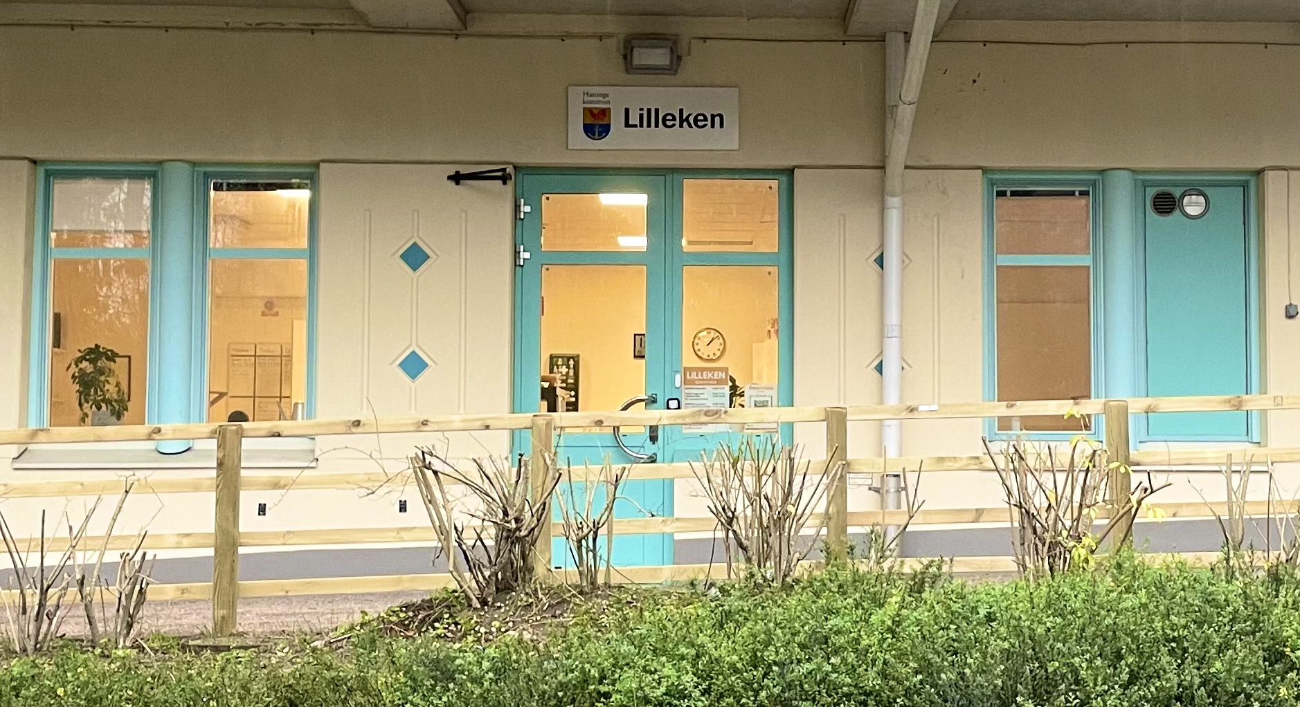 Entré till förskolan Lilleken i Haninge kommun. Byggnaden har ljus fasad med turkosa dörrar och fönsterkarmar. Ovanför dörren sitter en skylt med Haninge kommuns logotyp och texten ”Lilleken”. Framför entrén finns ett trästaket och några buskar.