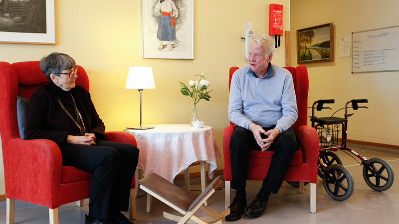 En äldre kvinna och en äldre man sitter i fåtöljer på ett äldreboende och pratar med varandra