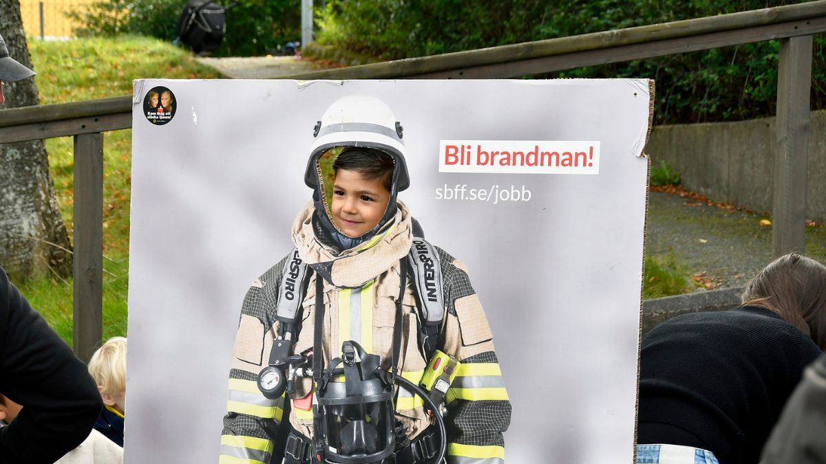 Pojke fotograferas medan han kikar i ett hål i en informationsskylt med brandmansklädsel.