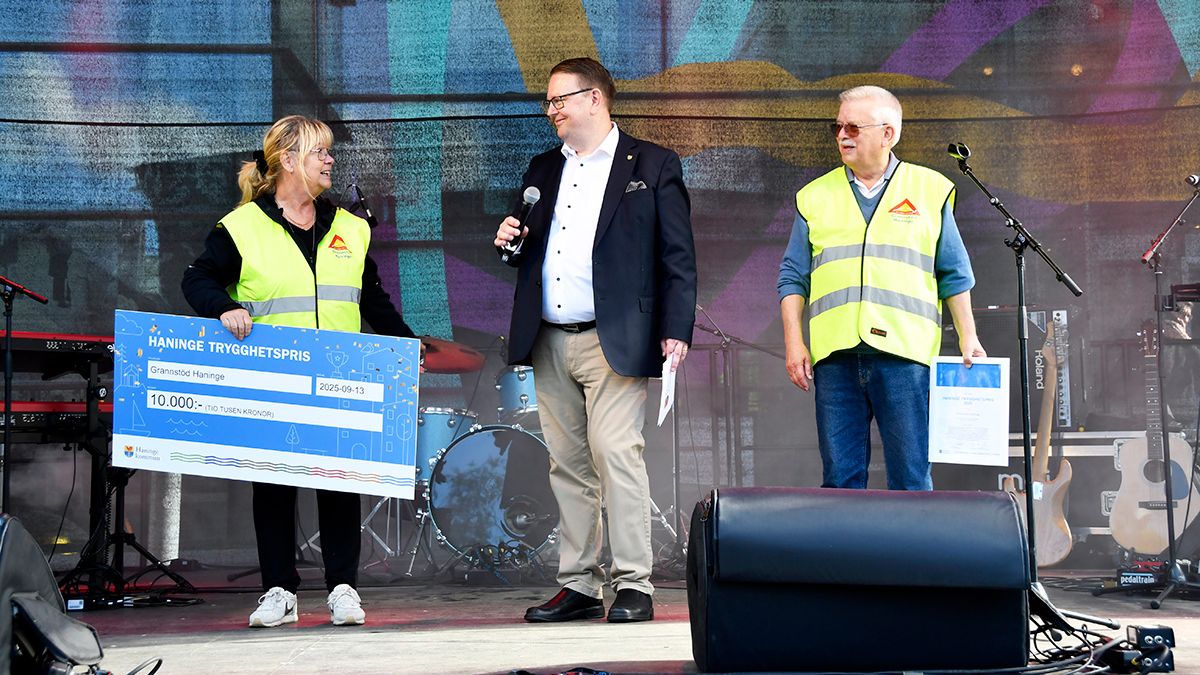 Tre personer står på en scen under ett evenemang. En person till vänster håller en stor symbolisk check för Haninge trygghetspris, den mittersta personen talar i mikrofon och en person till höger håller ett dokument. Instrument och scenutrustning syns i bakgrunden.
