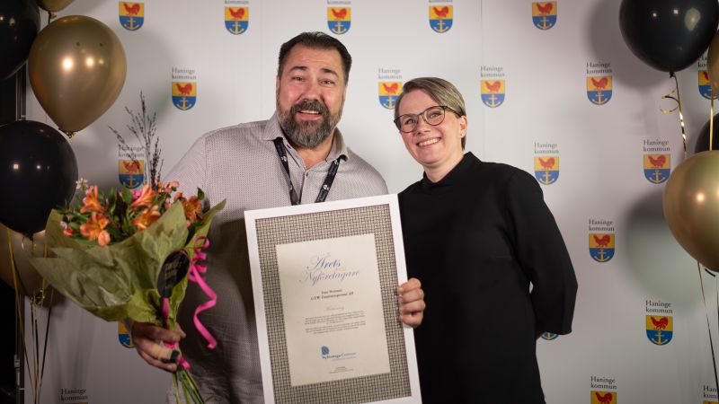 Två personer från GTW fotade med diplom och blommor, balloonger på sidorna och Haningeemblem  i  bakgrunden